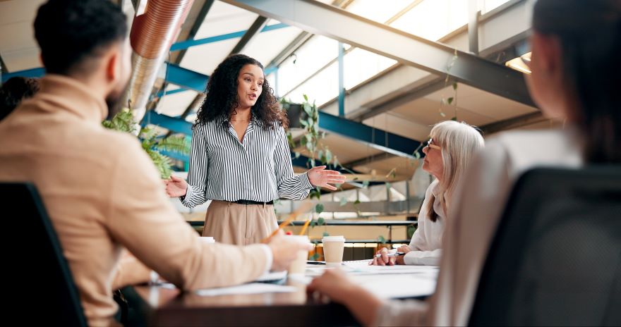 Marketing Campaigns June Article Image A woman in a striped shirt presents expressively in a bright meeting room, surrounded by attentive colleagues engaged in discussion.