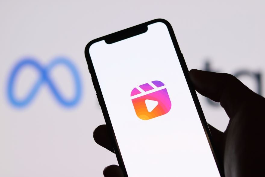 Meta Reels A hand holds a smartphone displaying the Instagram Reels icon with a blurred Meta logo in the background.
