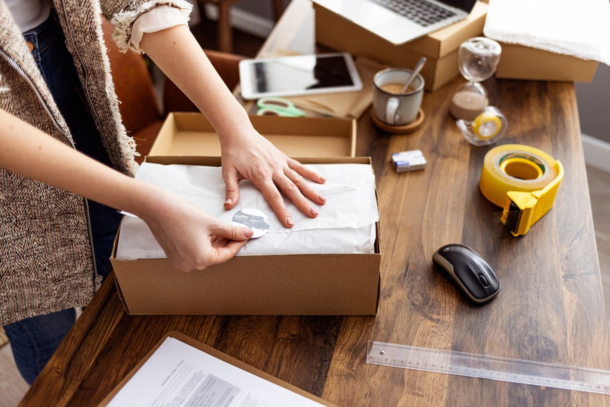 Young woman selling products online and packaging goods for shipping. Women, owener of small business packing product in boxes, preparing it for delivery. Person packaging a product on a table