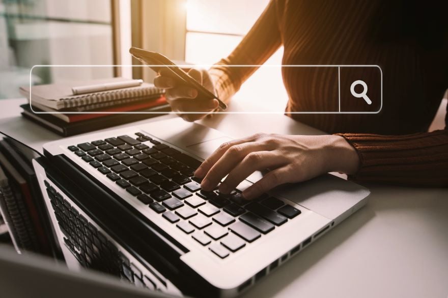 Hands typing on a keyboard while holding a phone with a search bar graphic on top